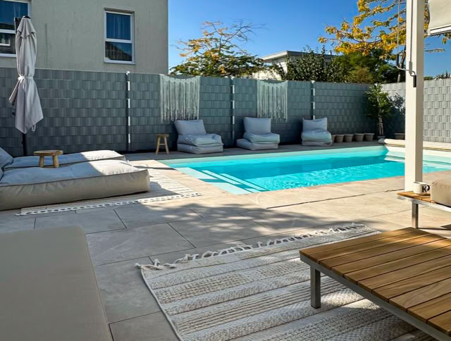 Ein moderner Außenbereich mit einem Pool, umgeben von Liegestühlen und Sonnenschirmen, sowie einem Holztisch auf einer Terrasse.