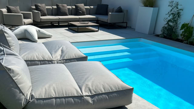 Sonnenliegen und ein Sofa mit Kissen neben einem blauen Pool auf einer Terrasse.