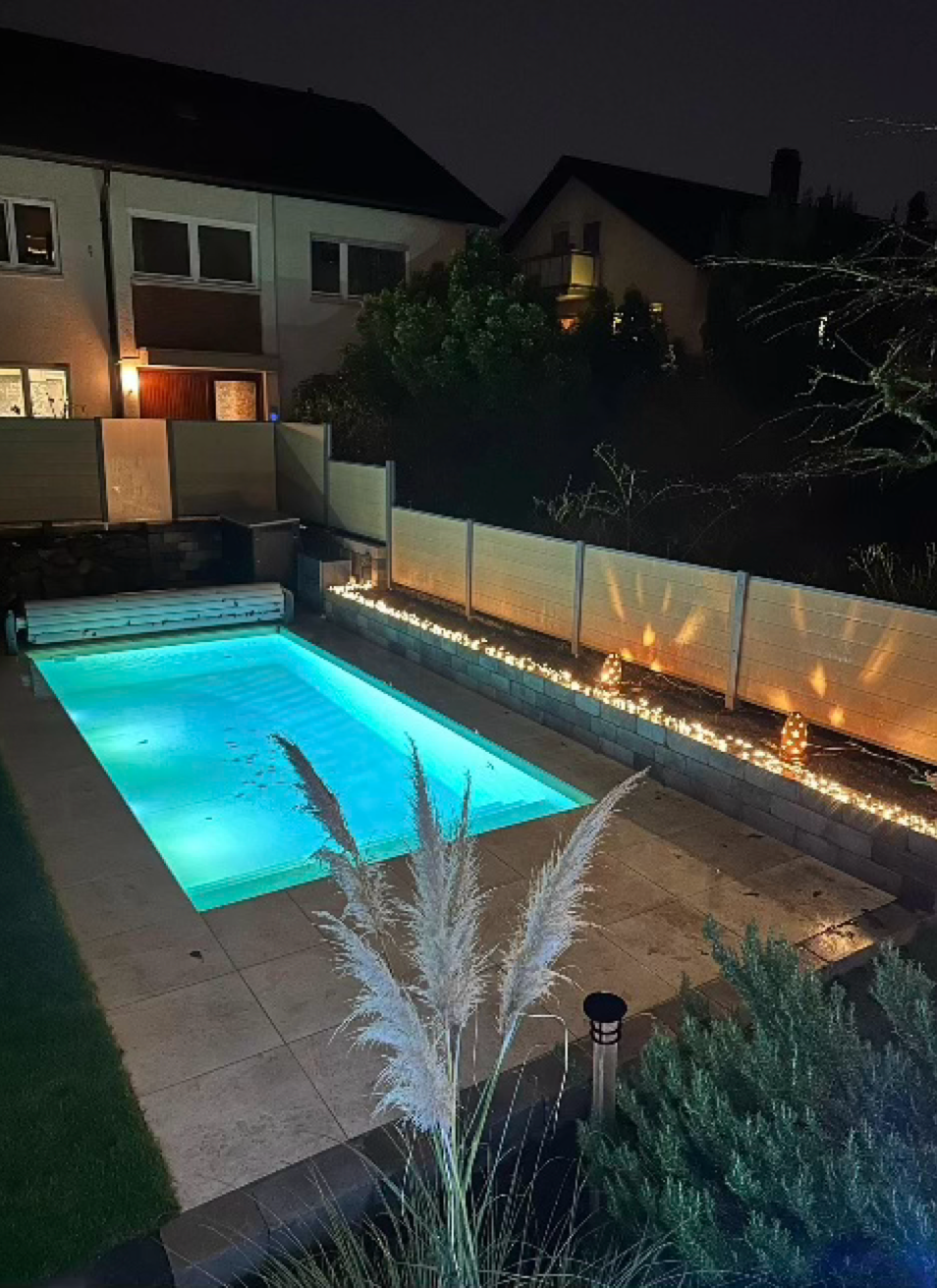 Beleuchteter Swimmingpool im Freien bei Nacht, umgeben von einem Zaun und Pflanzen, mit Häusern im Hintergrund.