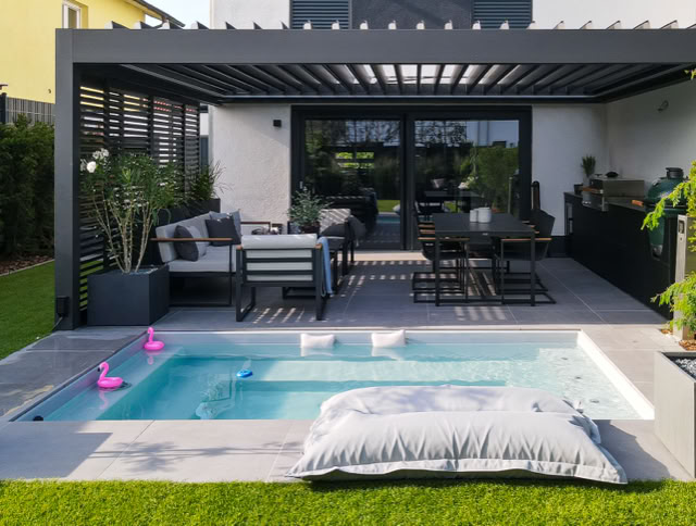 Moderne Terrasse mit Pergola, Sitzbereich, Esstisch und kleinem Pool im Vordergrund, umgeben von Rasen und Pflanzen.